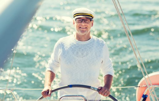 sailing, age, tourism, travel and people concept - happy senior man in captain hat on steering wheel and navigating sail boat or yacht floating in sea